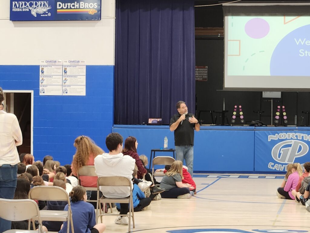 Presenter speaking to elementary students seated on a gym floor during a youth prevention assembly in an Oregon school gym.