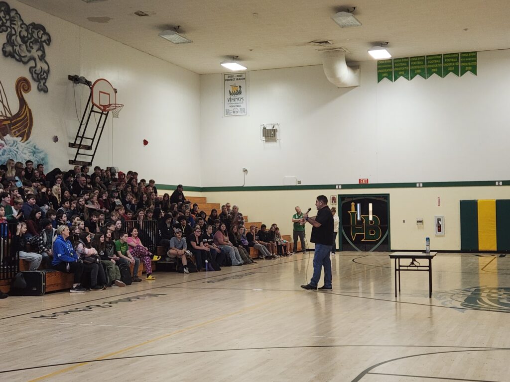Presenter speaking to a large group of middle school students seated on bleachers in a gym during a youth drug prevention assembly.