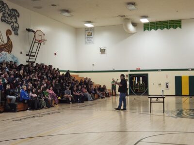 Presenter speaking to a large group of middle school students seated on bleachers in a gym during a youth drug prevention assembly.