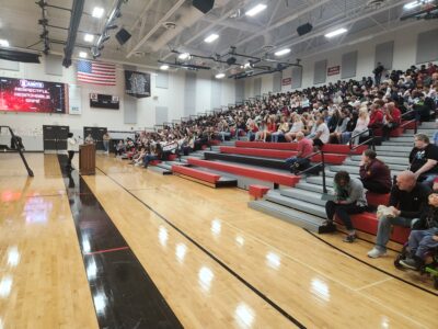 Ray Lozano presenting a youth drug prevention assembly to a full gym of students at Red Canyon High School, with students seated on bleachers listening attentively.