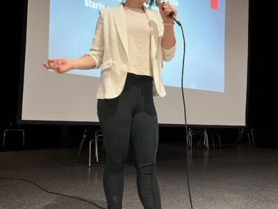 My daughter speaking into a microphone onstage during a youth drug prevention workshop, standing in front of a projected slide.