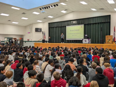 Ray Lozano speaking to a large group of middle school students seated on the floor in a school auditorium during a youth drug prevention assembly.