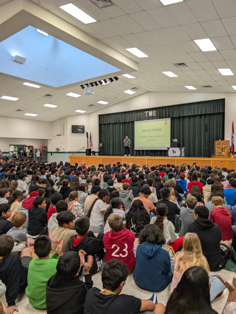 Ray Lozano speaking to a large group of middle school students seated on the floor in a school auditorium during a youth drug prevention assembly.