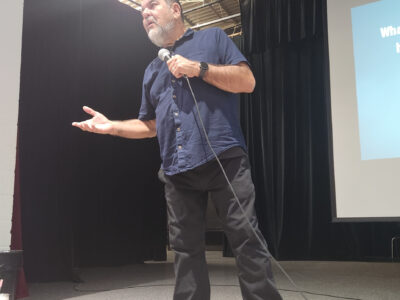 A presenter speaking onstage with a microphone during a youth drug prevention assembly, addressing students about decision-making and substance-use risks.