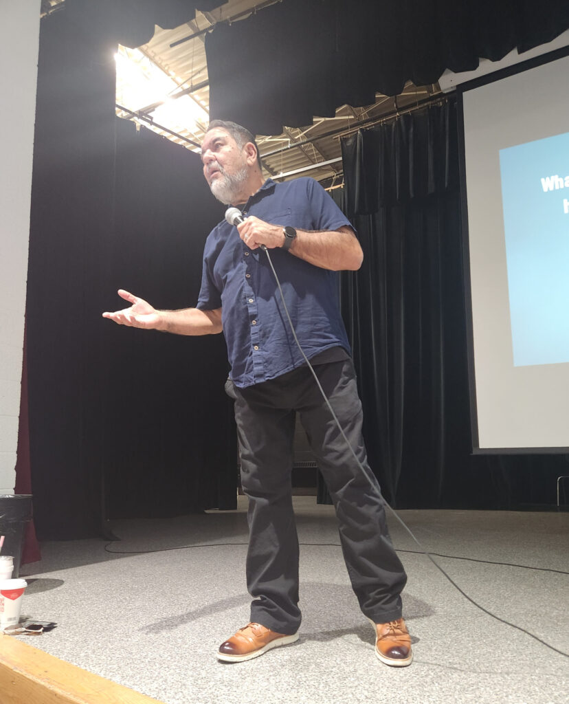 A presenter speaking onstage with a microphone during a youth drug prevention assembly, addressing students about decision-making and substance-use risks.