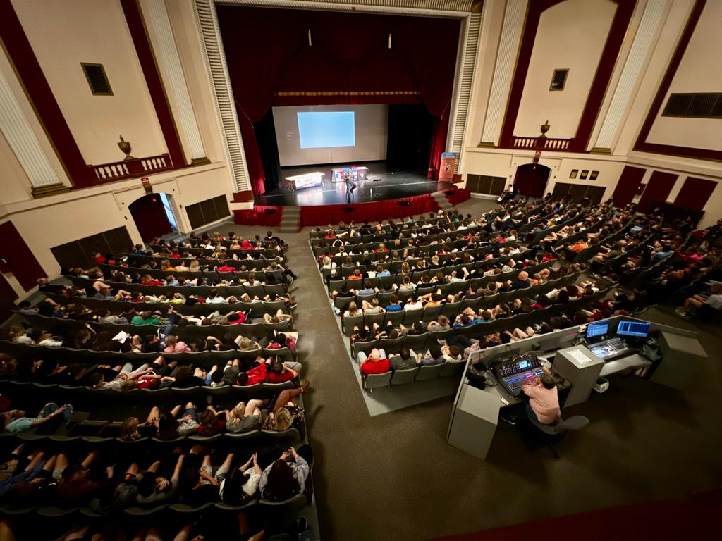 Large auditorium filled with students attending a school-wide youth drug prevention presentation led by a professional prevention speaker.