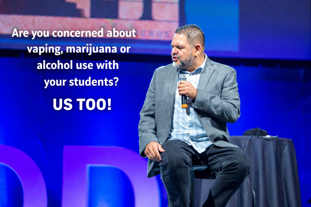 Ray Lozano, a youth prevention educator, sits on a stage holding a microphone during a professional presentation, with on-screen text addressing concerns about vaping, marijuana, and alcohol use among students.