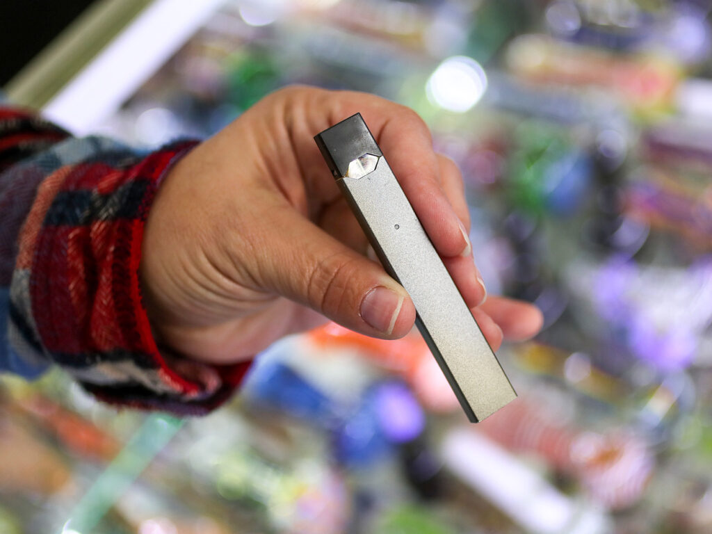 Hand holding a silver JUUL vape device in a store setting, highlighting youth access and vaping risks.